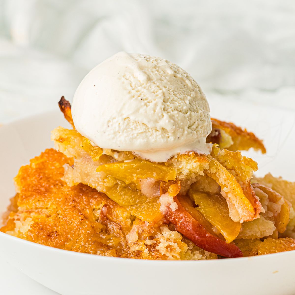 Old fashioned peach cobbler served in a white bowl is vanilla ice cream on top